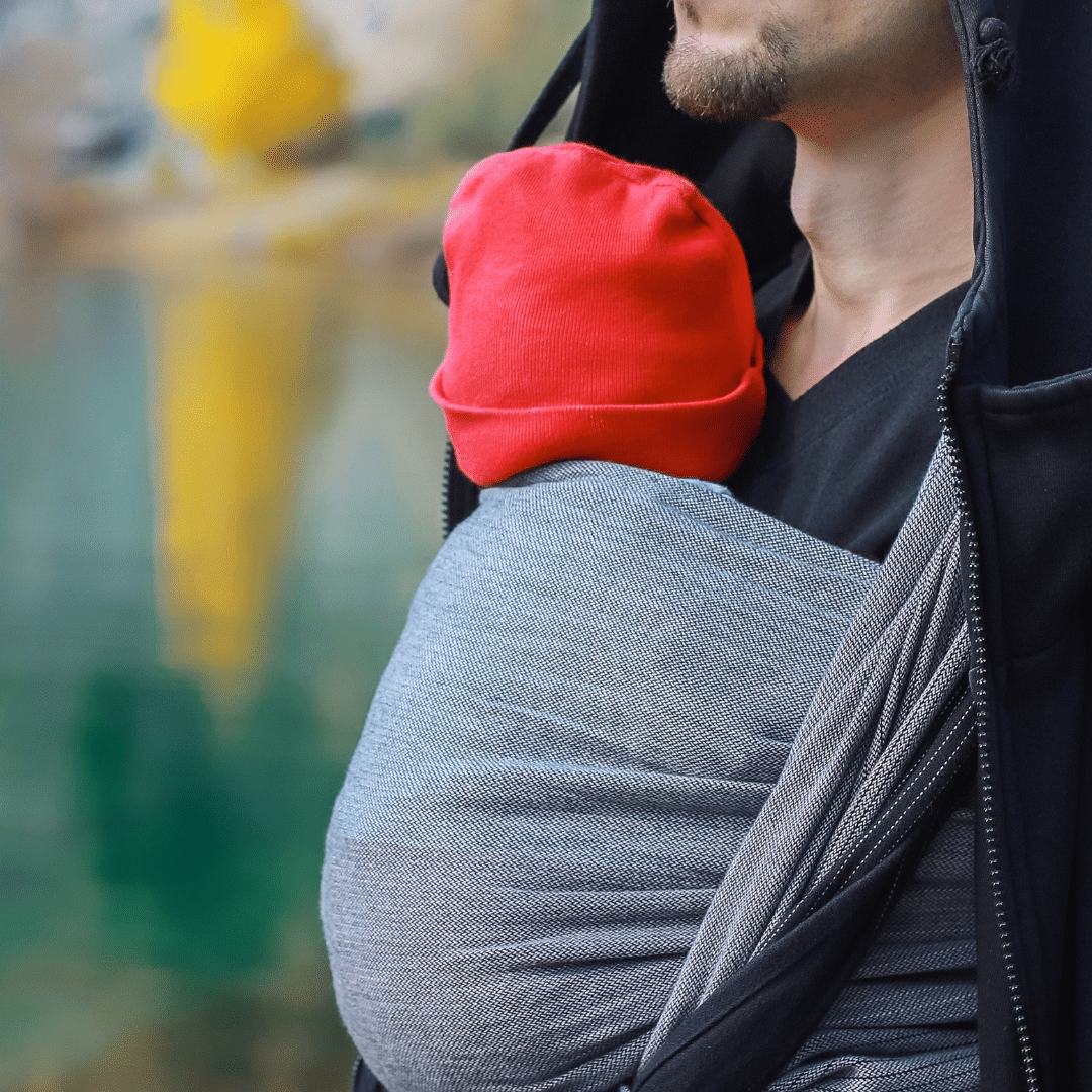 Babywearing using a woven wrap. Baby wears a red hat and is carried by a male